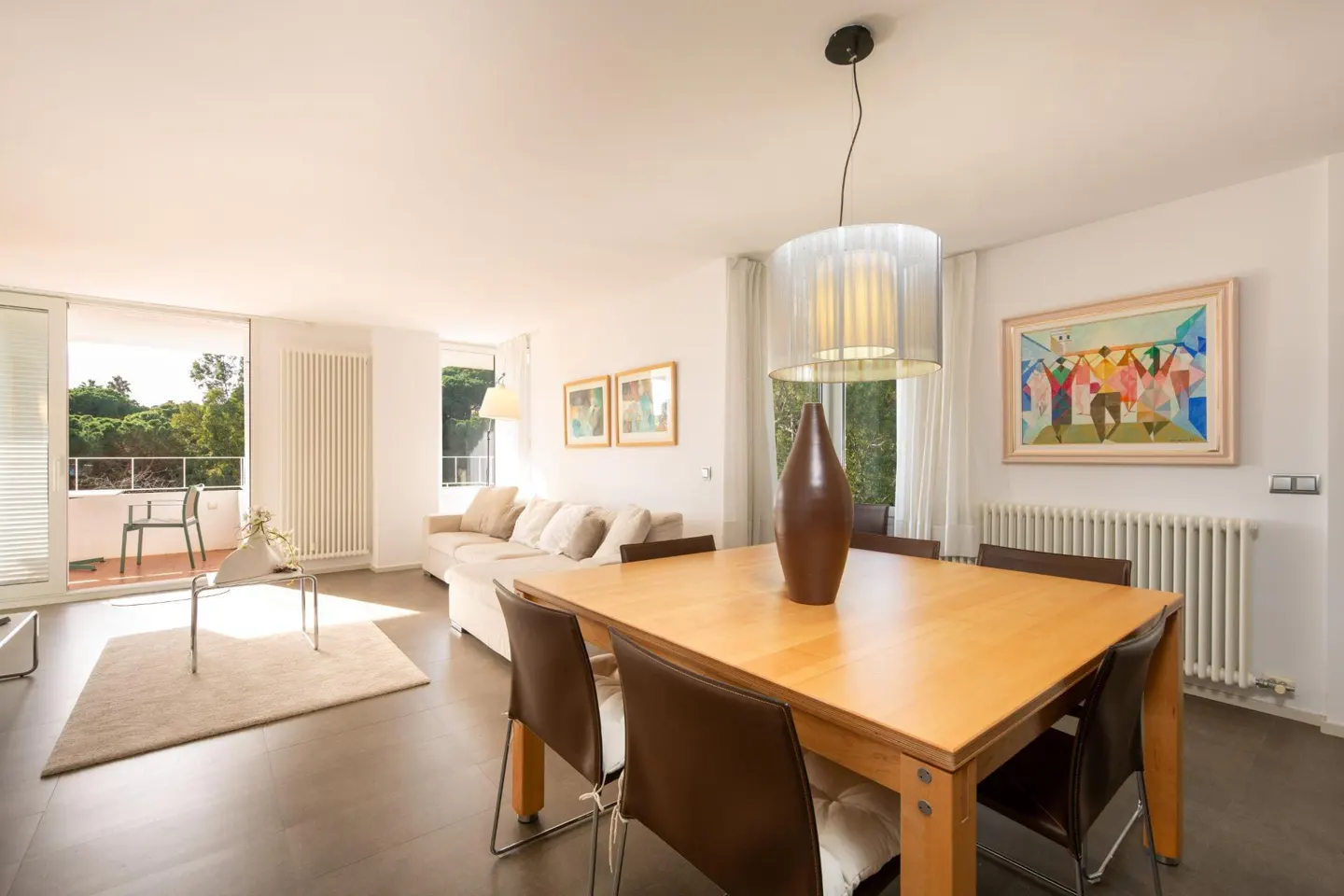 Bright living room with a wooden dining table, brown chairs, and a large vase. A beige sofa and balcony are visible in the background.