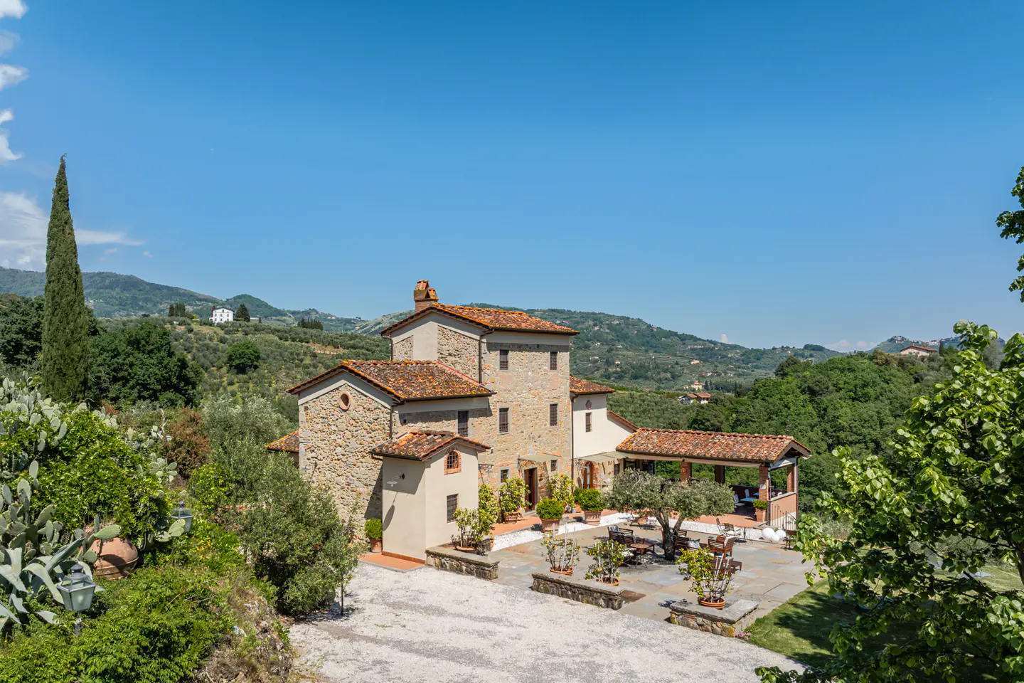 Exterior view of a stone Tuscan villa with a red tile roof, patio, and green trees under a clear blue sky.