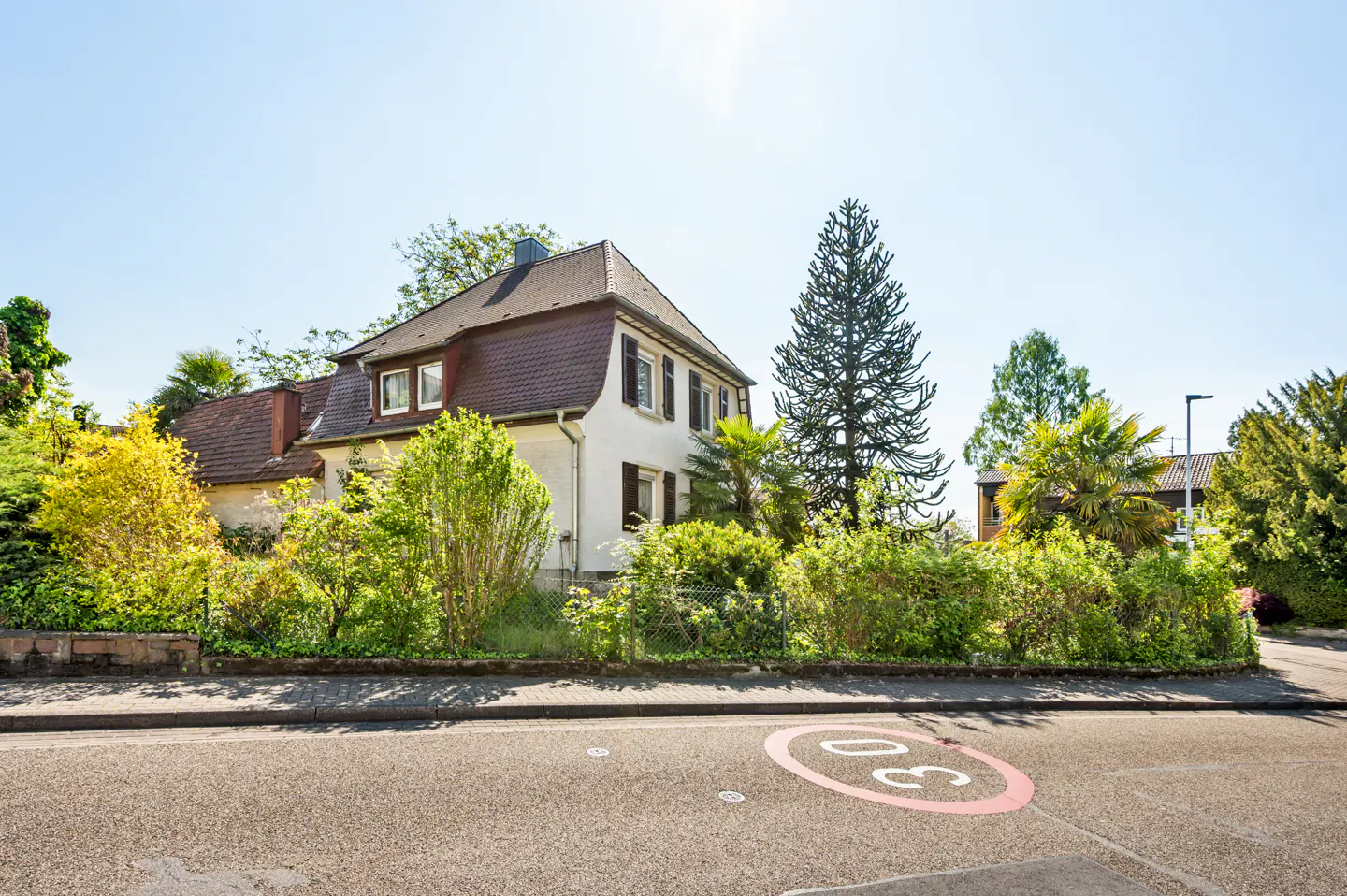 Exterior view of a two-story house with a brown roof, white walls, and dark shutters, surrounded by lush greenery. A "30" speed limit sign is painted on the road.