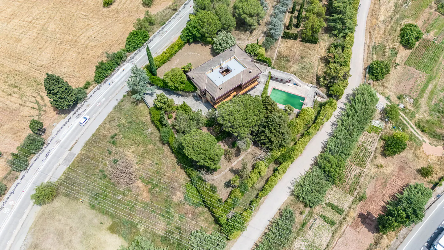 Aerial view of a house with a green pool, surrounded by trees and fields, situated between two roads.