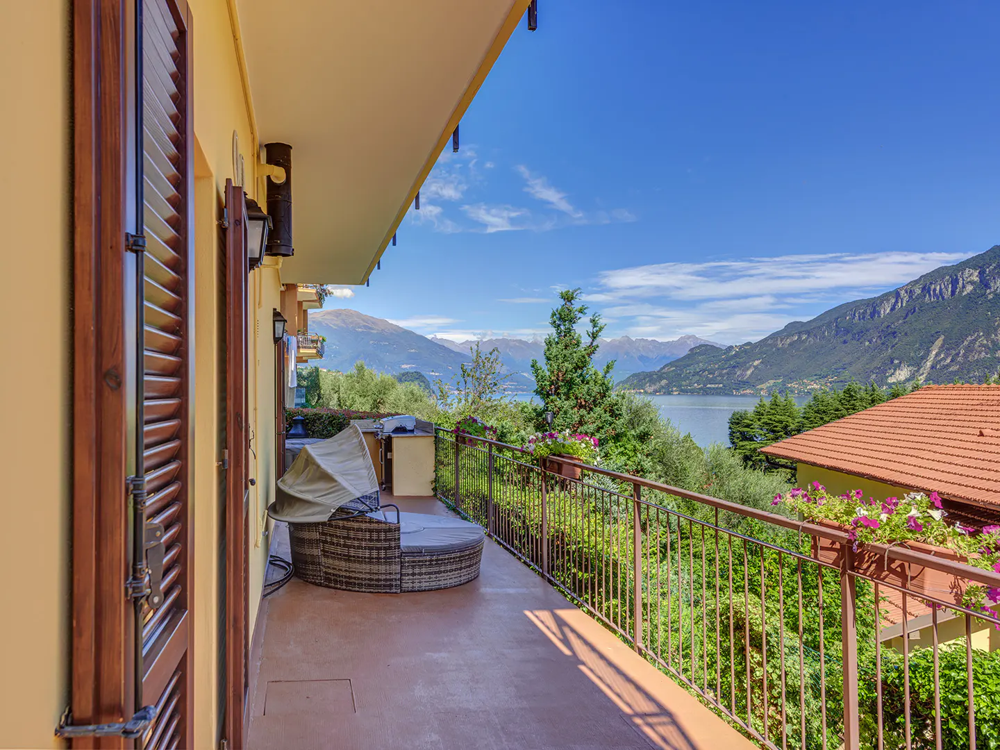 Balcony view of Lake Como, Italy. Wicker furniture sits on the patio, with mountains and blue sky in the background.