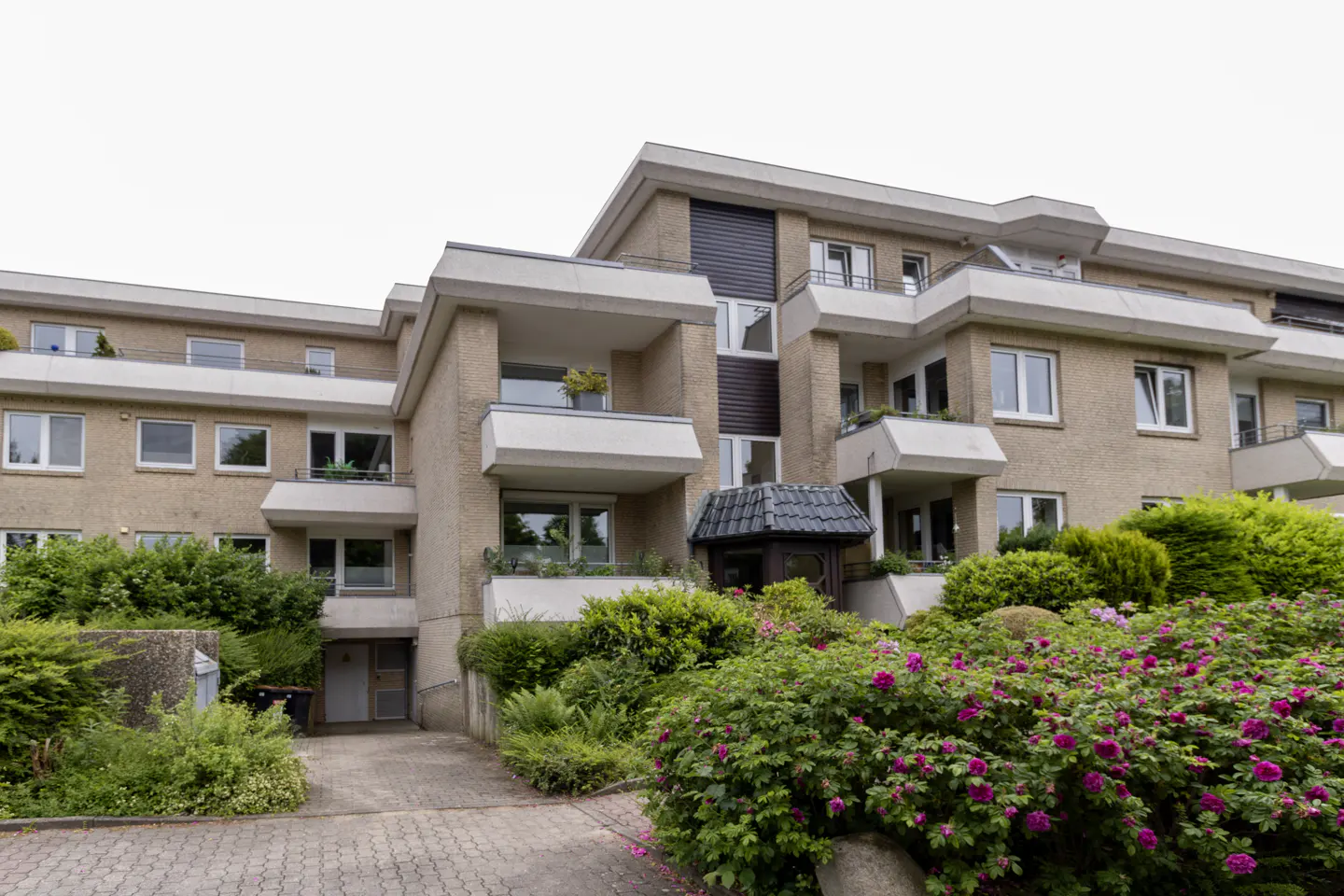 A multi-story beige apartment building with balconies, surrounded by lush green bushes and blooming pink rose bushes.