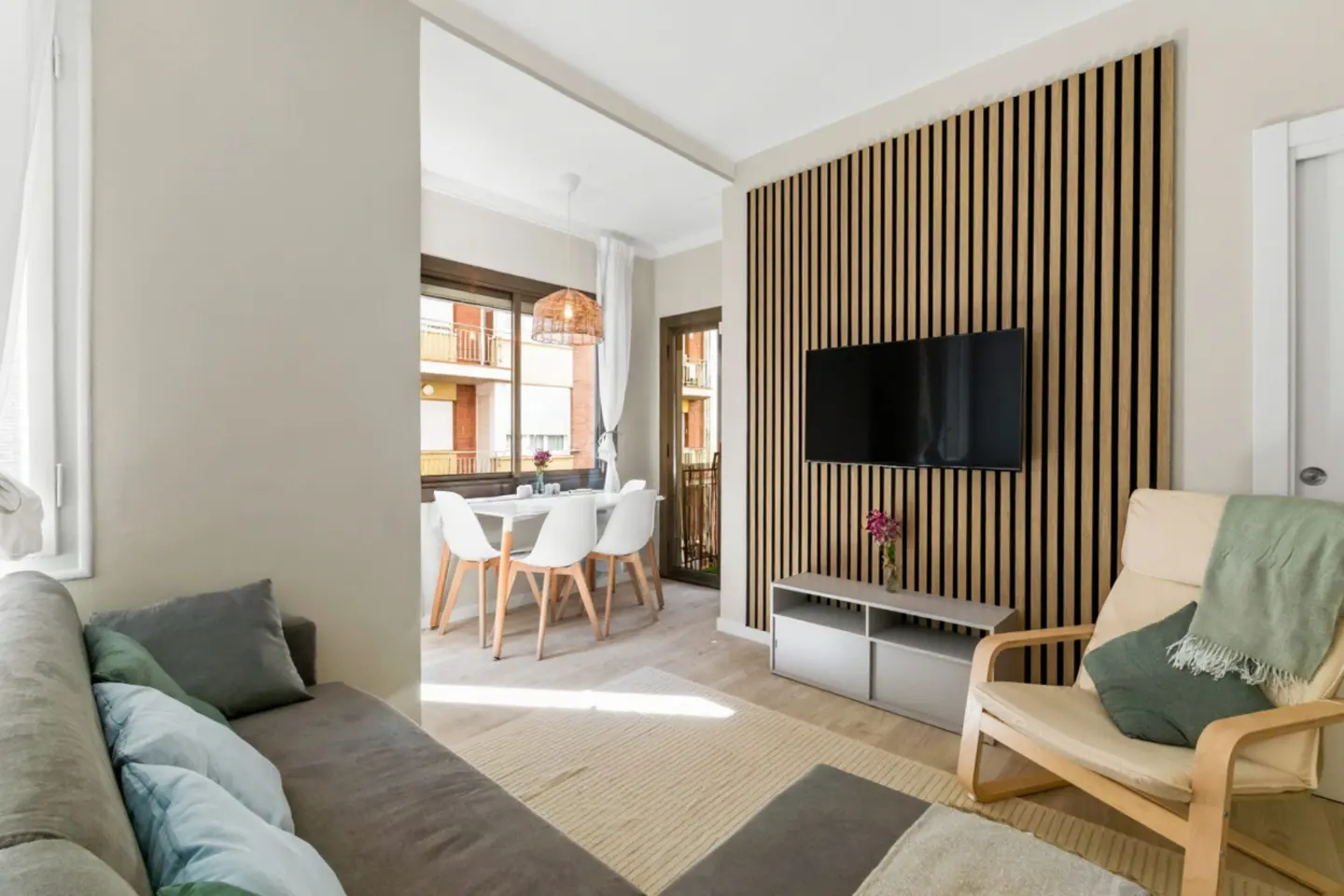 Living room with a gray sofa, beige armchair, and a dining area with white chairs and a table. A TV is mounted on a wooden slat wall.