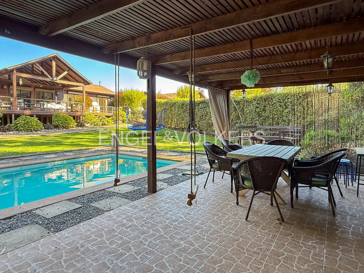 Covered patio with table, chairs, and swings overlooking a pool, lawn, and house.