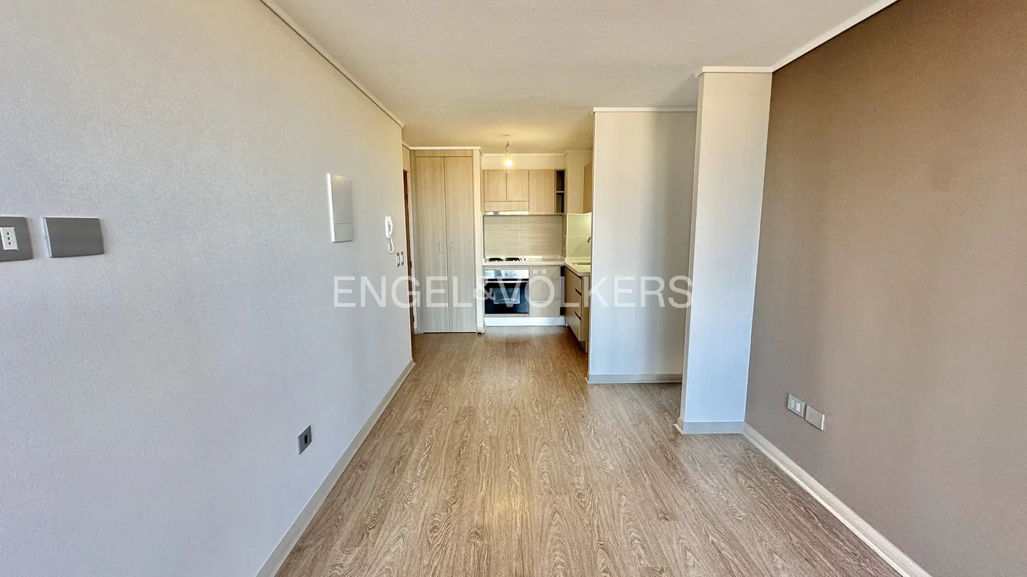 Apartment interior view with wood floors, white and beige walls, and a glimpse of the kitchen with light wood cabinets.