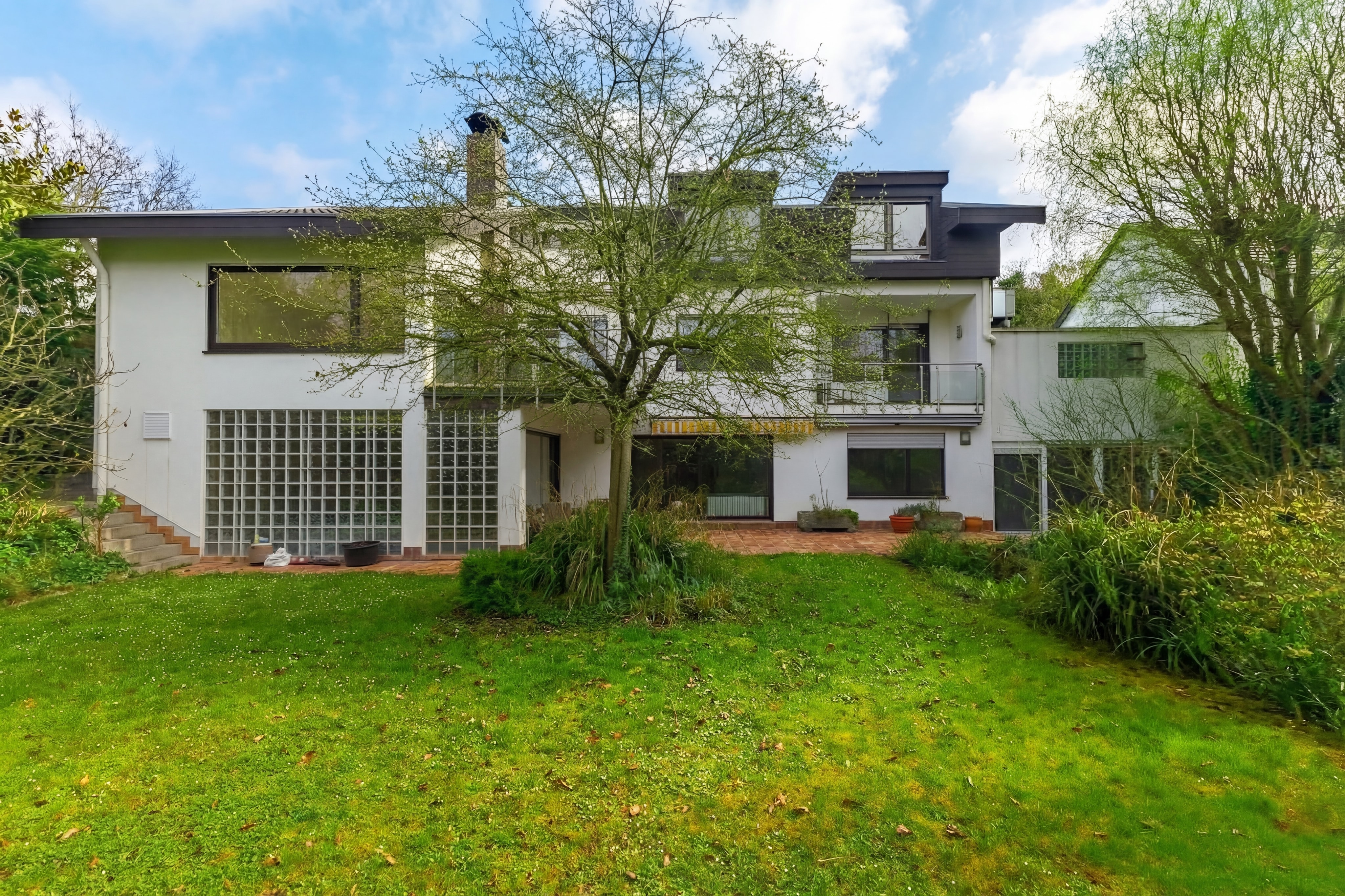 Modern white house with glass block walls, a balcony, and a large tree in the center of a green backyard lawn. [AI-generated text]