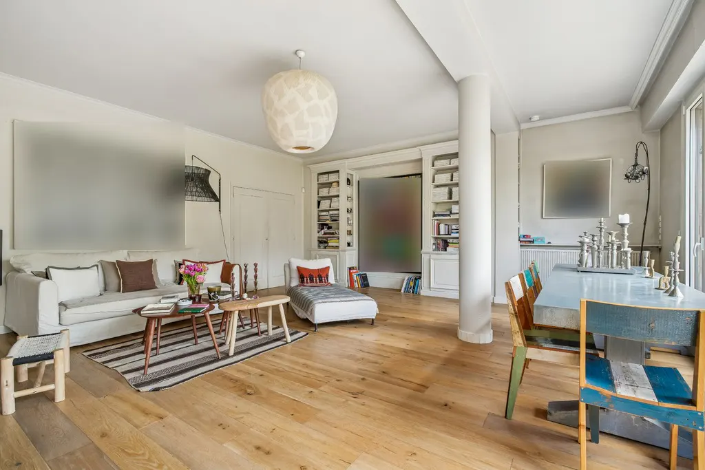 Bright living room with hardwood floors, white sofa, striped rug, and a dining table with colorful chairs. Bookshelves and a large white pendant light add character.