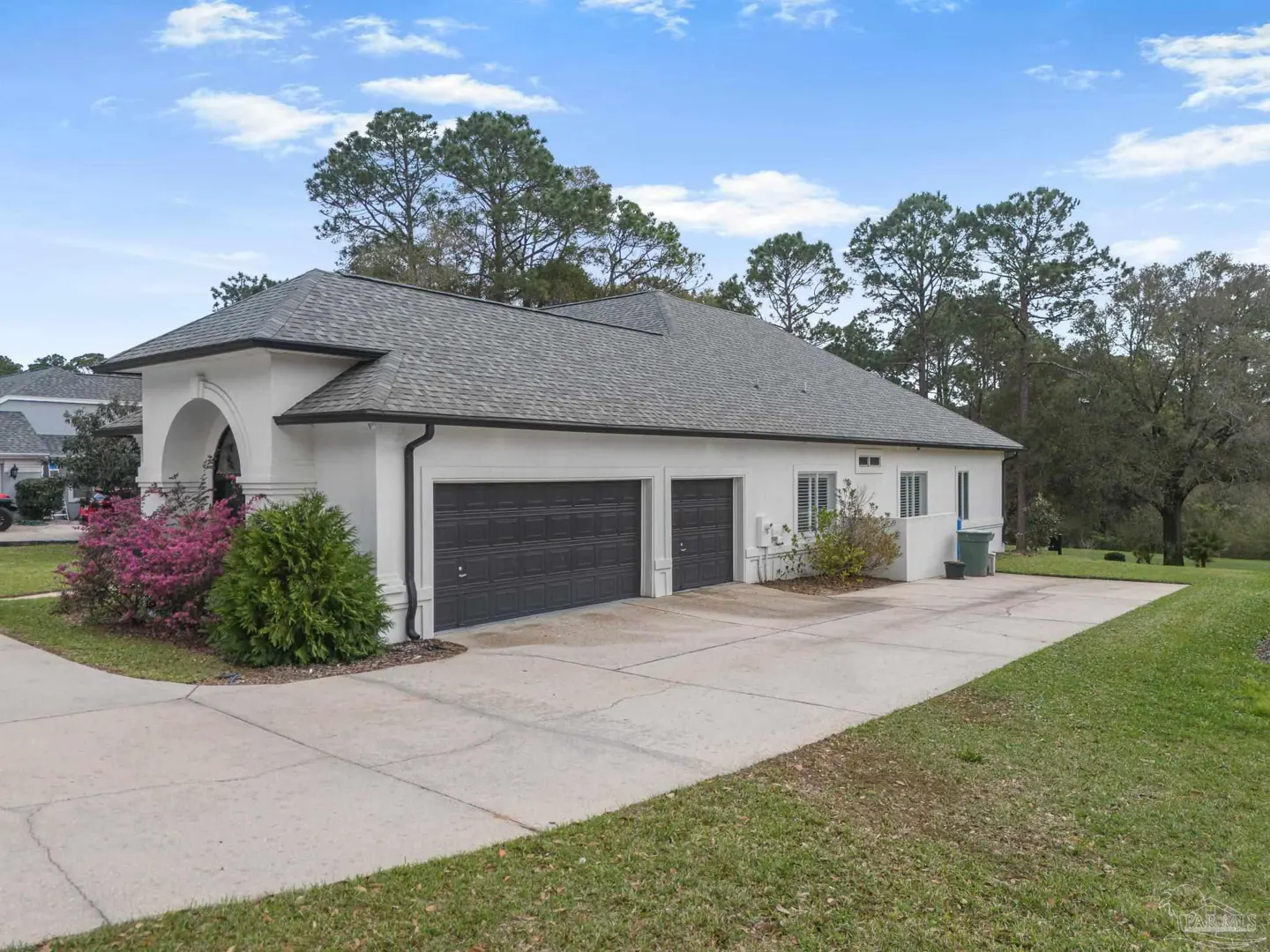White house with a gray roof, two dark garage doors, a concrete driveway, and landscaping against a backdrop of trees.