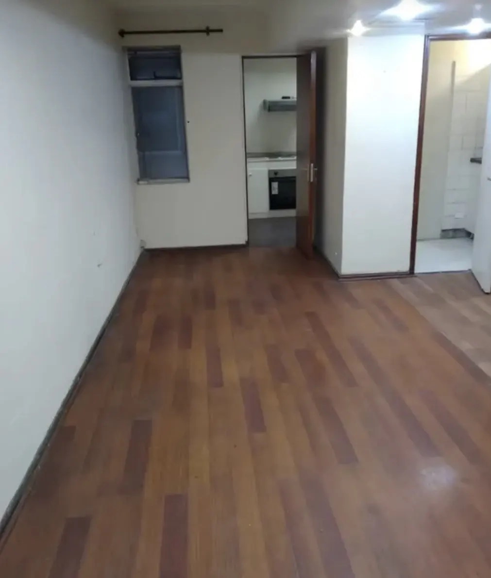 An empty room with wood floors, white walls, and a window. An open doorway leads to a kitchen with white cabinets and appliances.