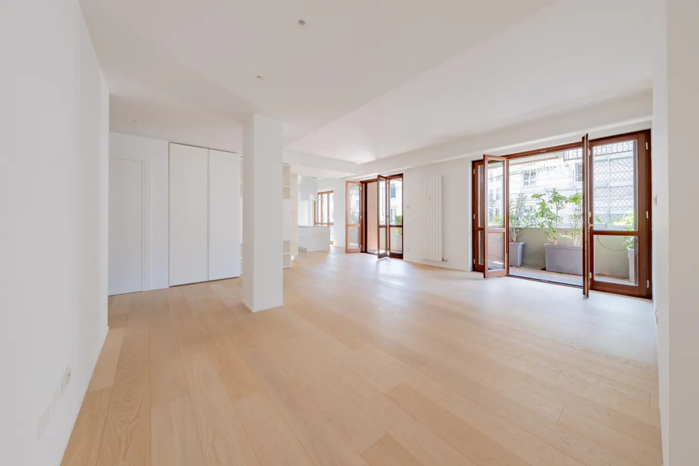 Bright, empty room with light wood floors, white walls, and open wood-framed doors to a balcony with plants.