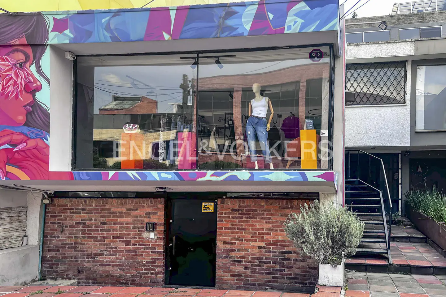Storefront with a large window displaying a mannequin in jeans and a white top. A colorful mural adorns the building's side. Brick facade and black door below.