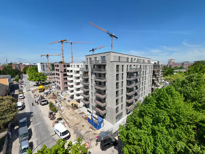 View of new apartment buildings under construction, with cranes, trees, and a street with cars.