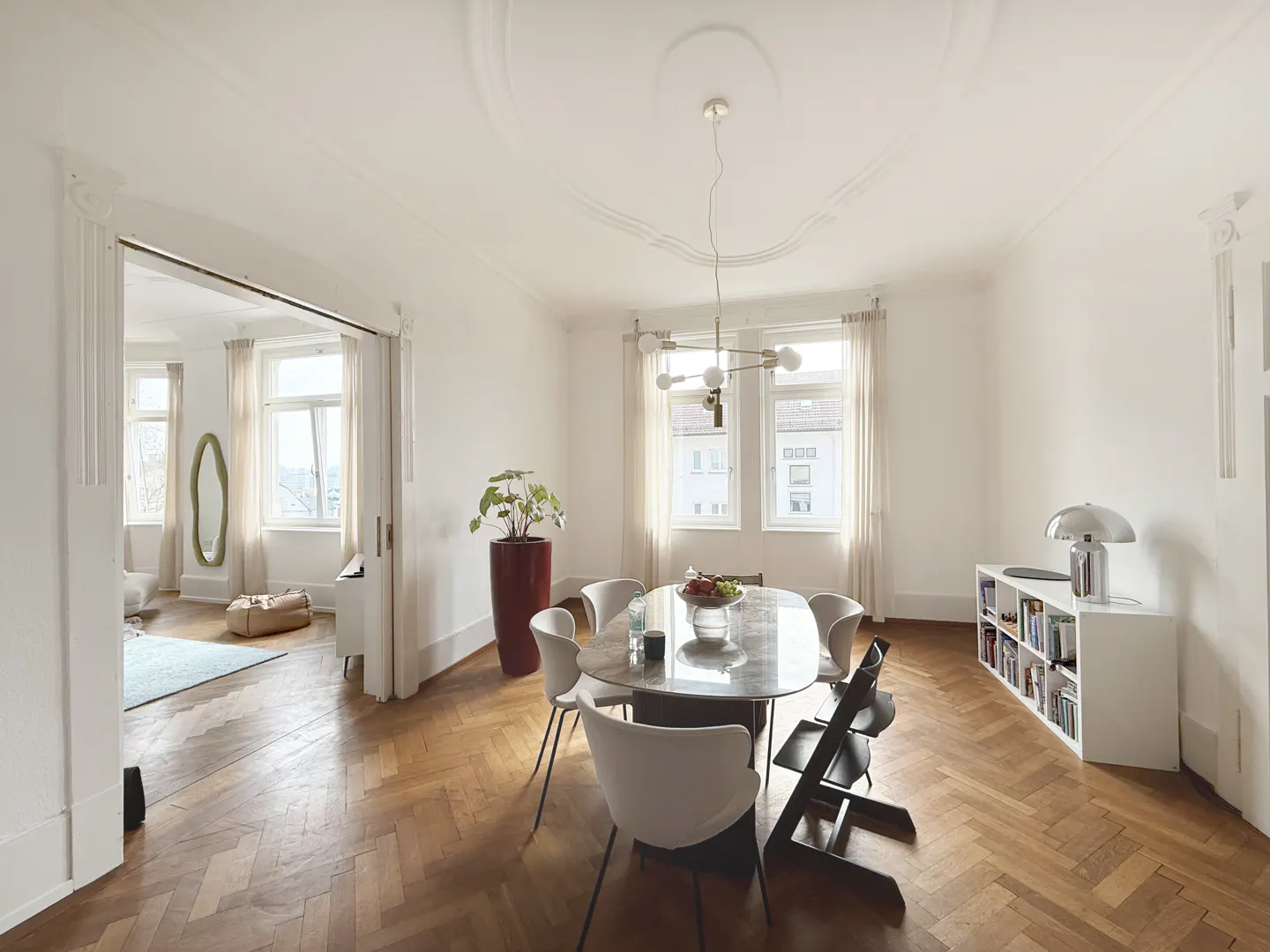 Bright dining room with herringbone wood floors, white walls, and a marble table with white chairs. A red planter and white bookshelf add color.