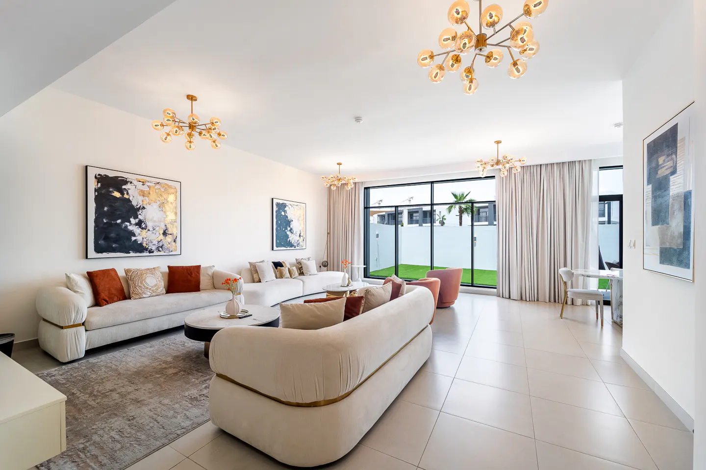 Bright, modern living room with white sofas, abstract art, and gold chandeliers. Large windows overlook a green lawn.