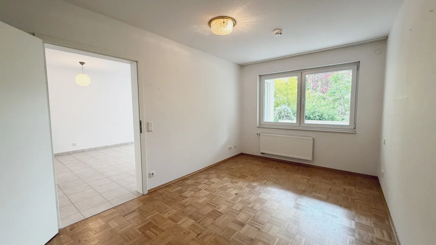 Bright, empty room with parquet floor, white walls, and a window overlooking greenery. A doorway leads to another room with tile flooring.