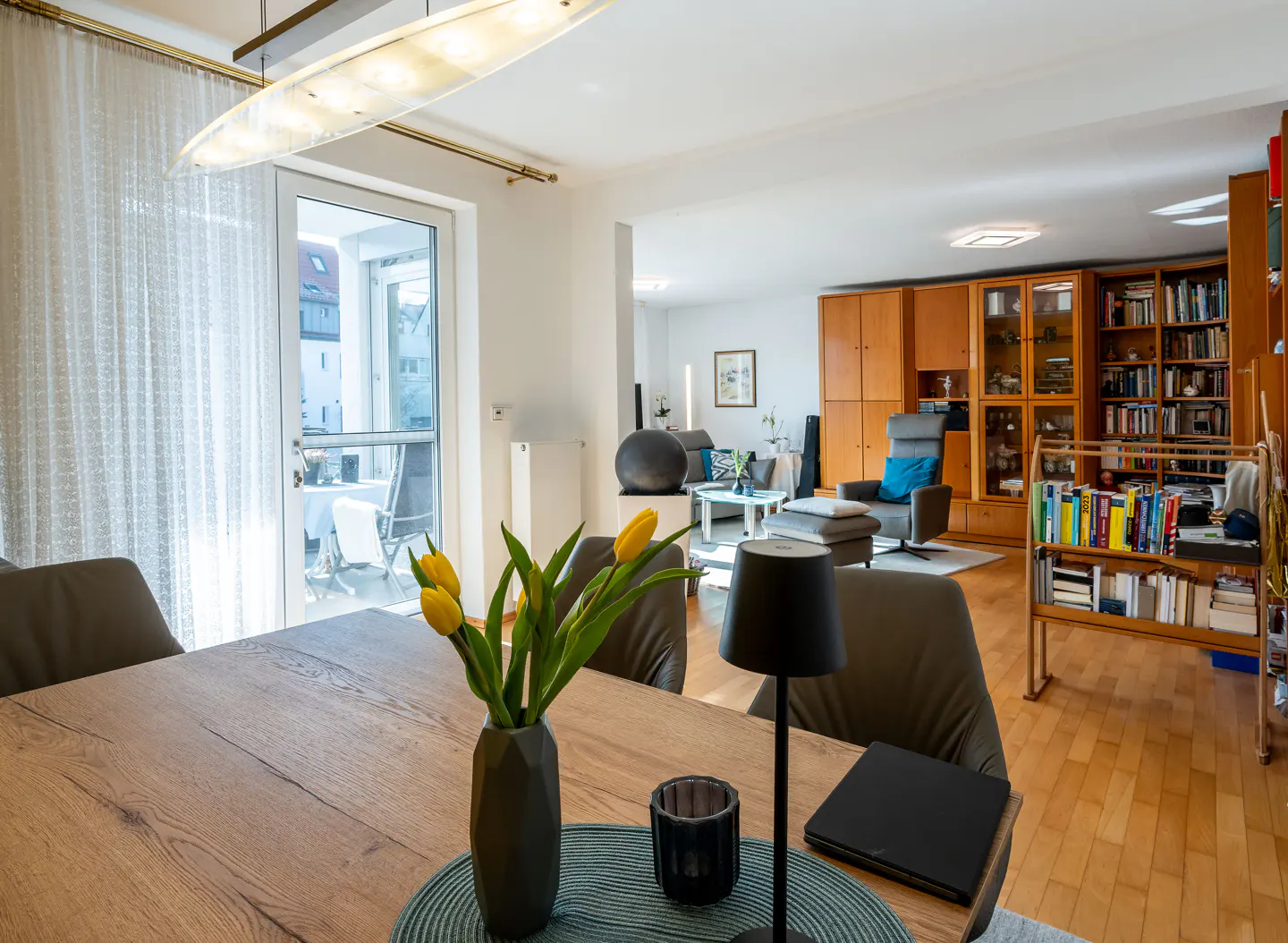 Open-concept living space with dining table, chairs, and yellow tulips. Balcony visible through sheer curtains.