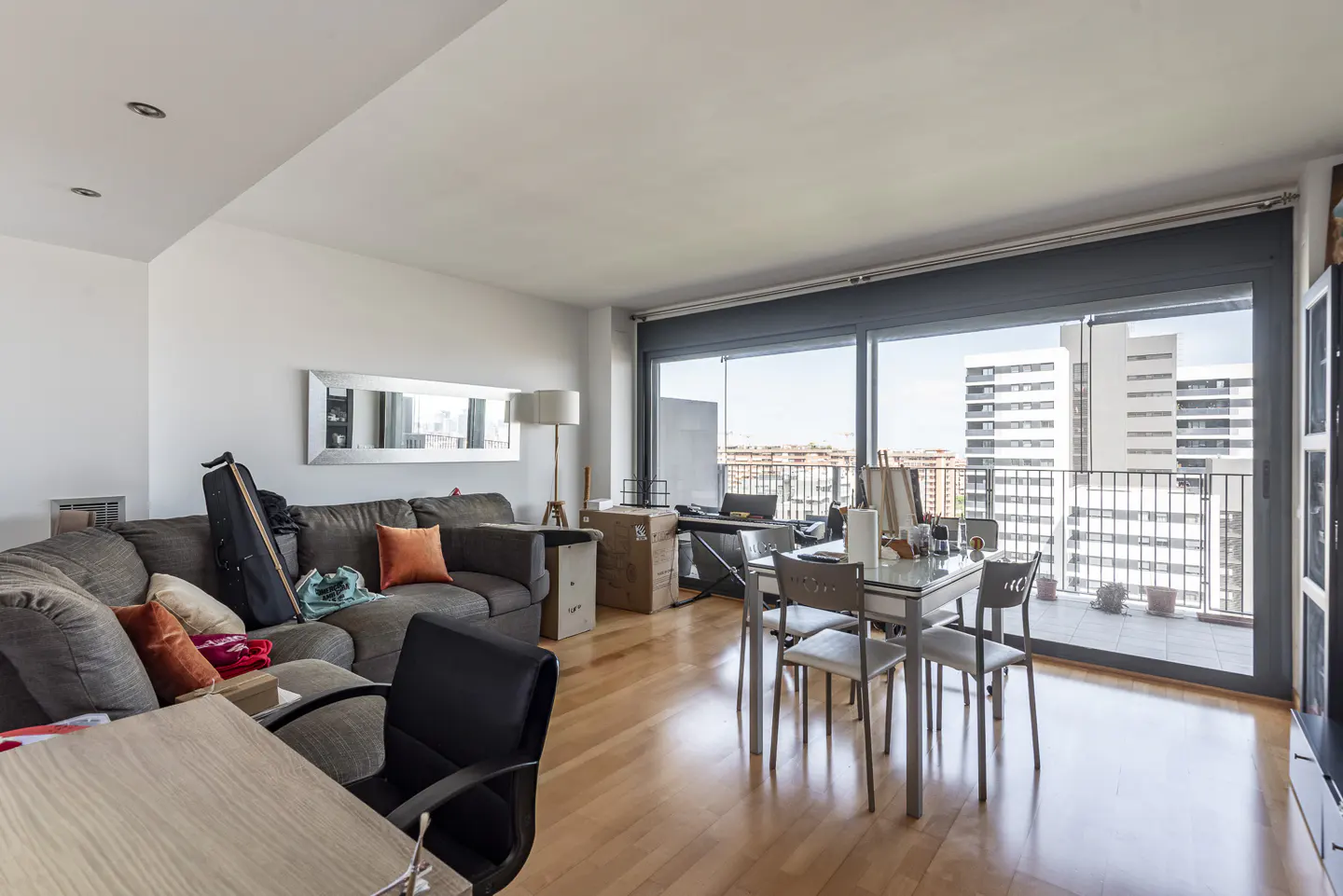 Bright living room with hardwood floors, a gray sectional sofa, a dining table with four chairs, and a large window with a city view.
