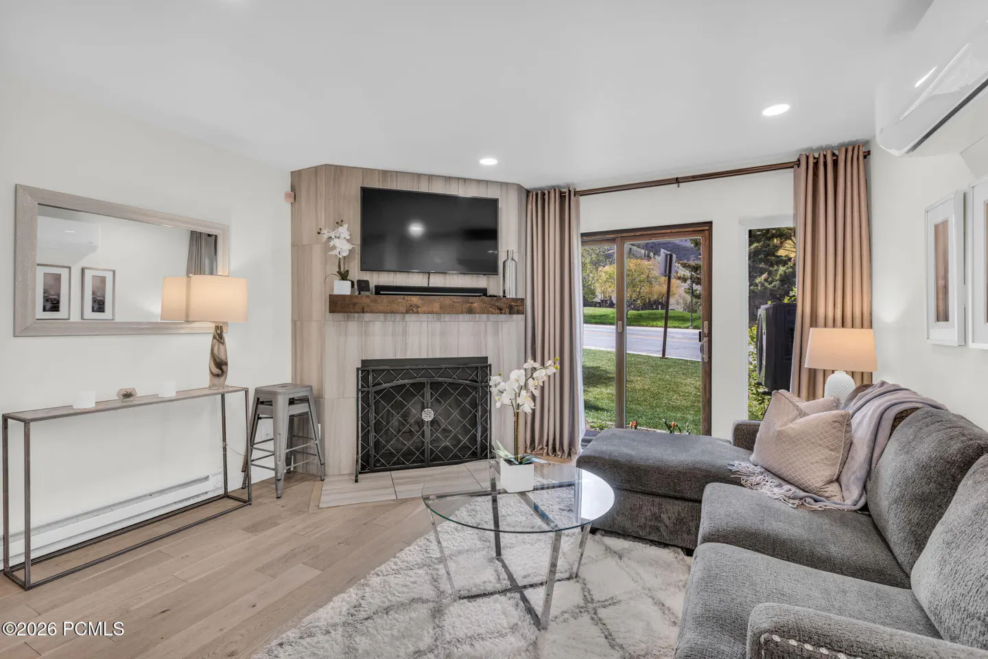 Living room with gray sofa, glass table, and fireplace with TV above. Sliding glass doors lead to a green lawn.