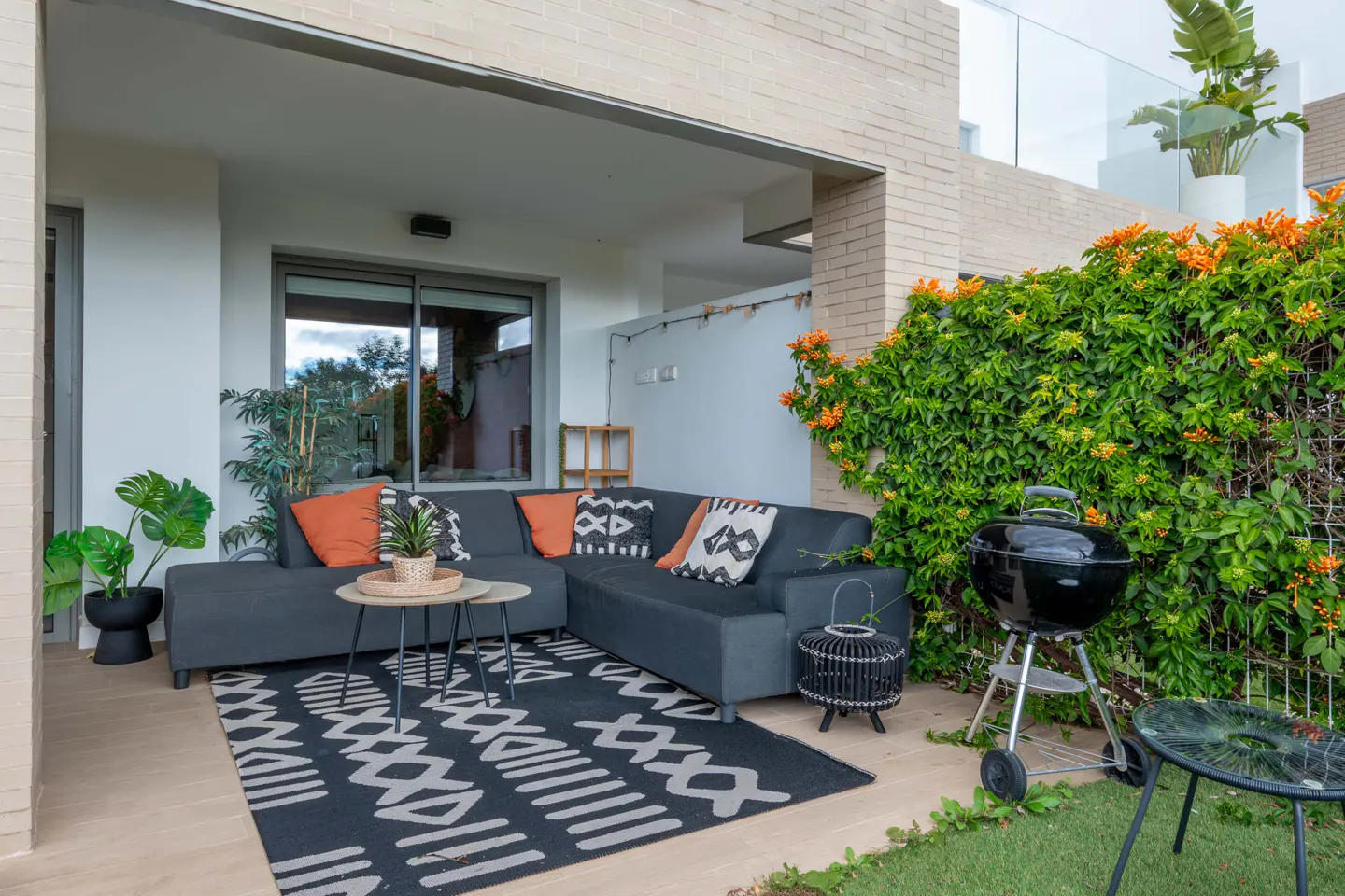 Outdoor patio with a gray sectional sofa, patterned rug, grill, and lush greenery with orange flowers.