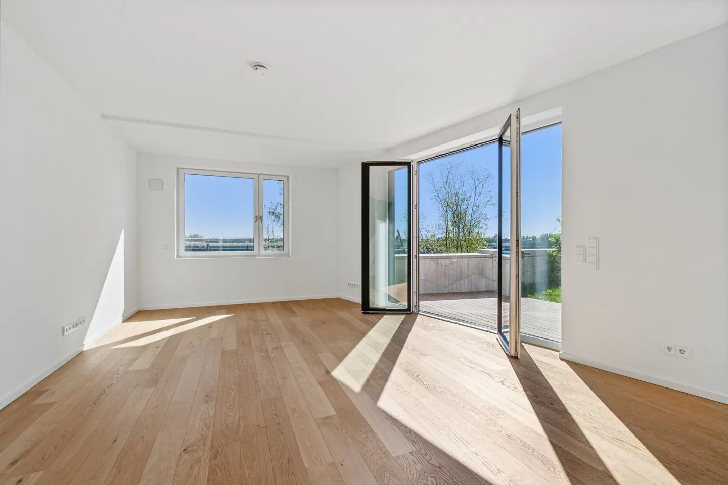 Bright, empty room with wood floors, white walls, and open glass doors to a balcony. A window shows a blue sky and green landscape.