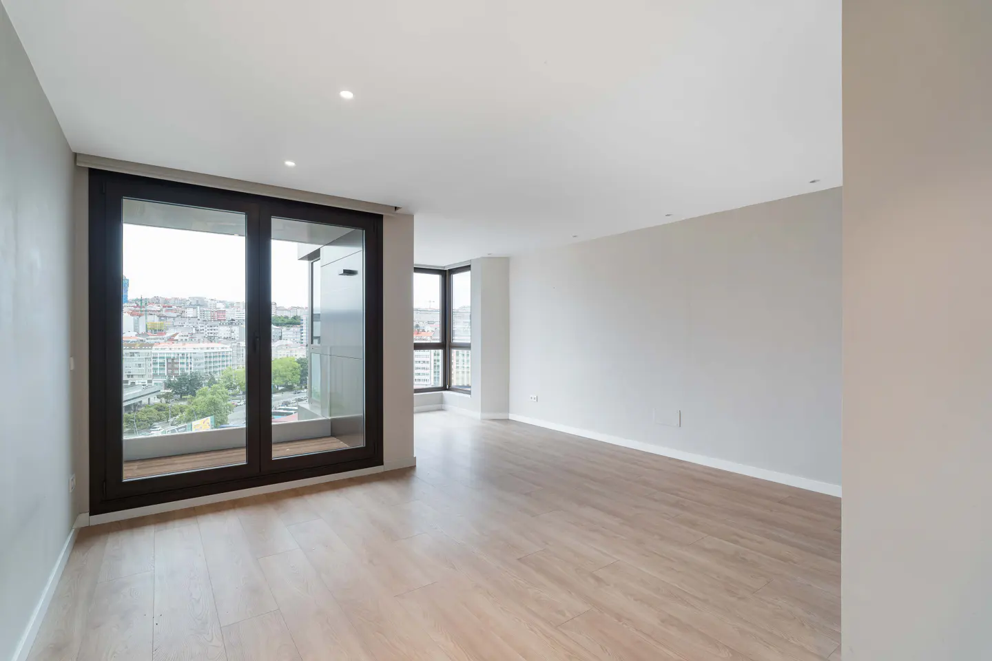 Bright, empty room with light wood floors, white walls, and dark-framed windows overlooking a cityscape.