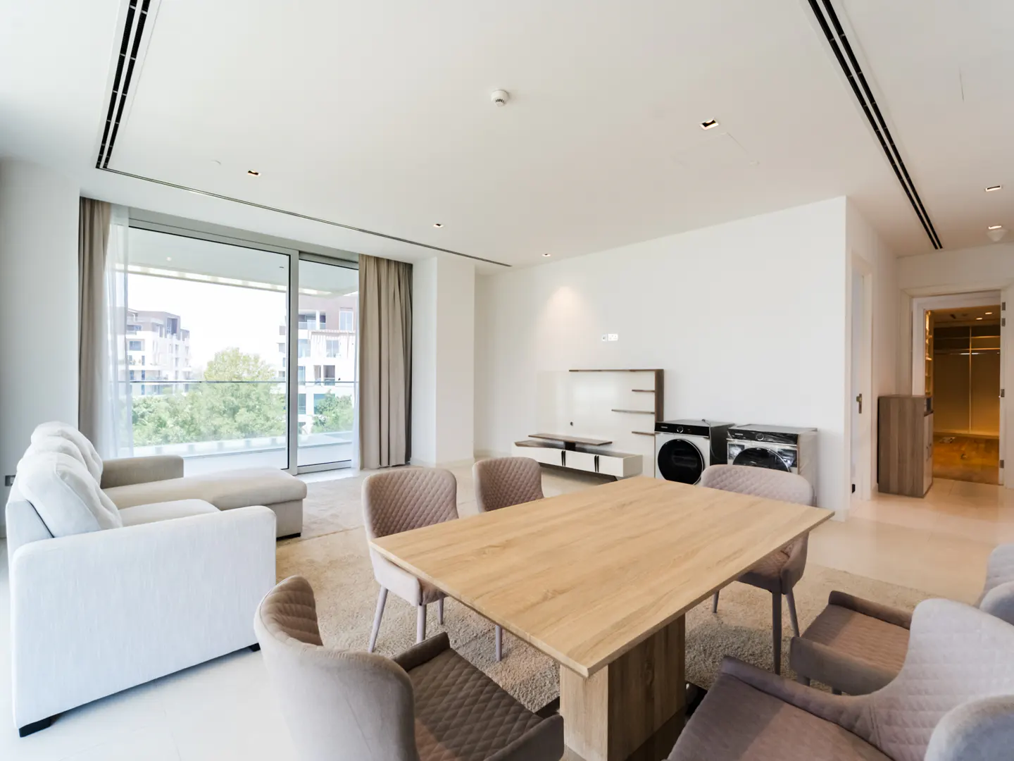 Modern open-plan living area with a white sofa, wooden dining table, quilted chairs, and floor-to-ceiling windows.