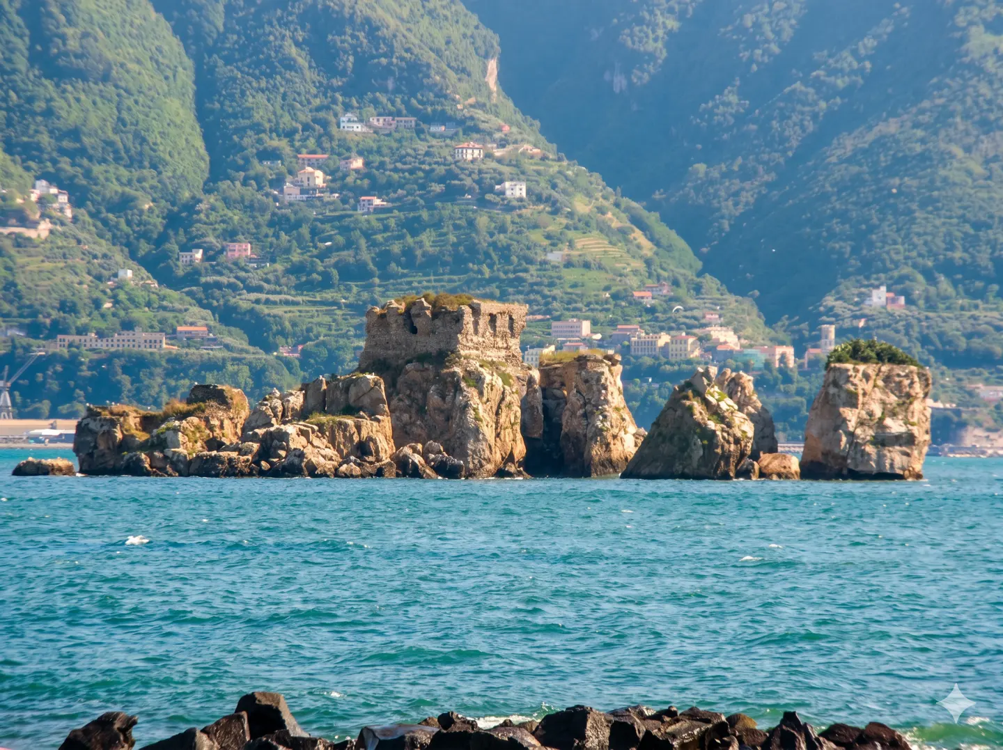 Jagged rock formations rise from turquoise sea waters, with a lush, mountainous coastal town in the background.