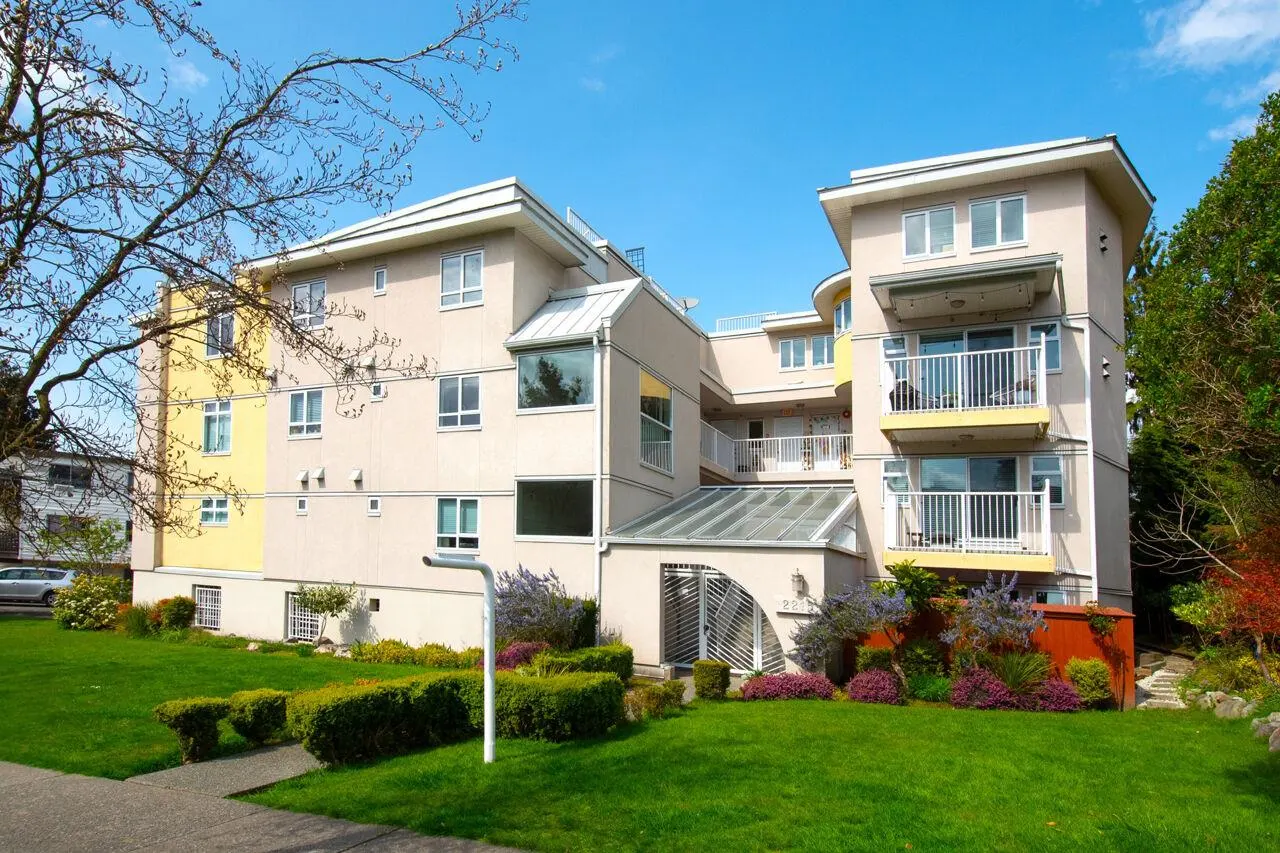 Exterior view of a multi-story apartment building with balconies, a green lawn, and a blue sky.