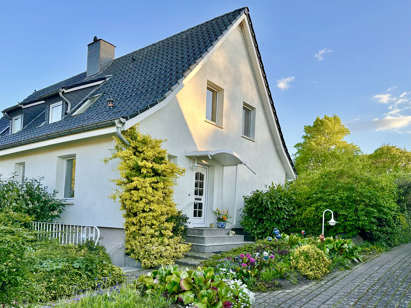 A white house with a dark roof, green bushes, and a stone walkway under a blue sky.