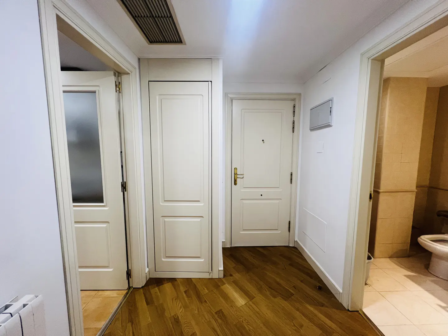 Hallway with wood floors, white walls, and three white doors. One door leads to a bathroom with a toilet.