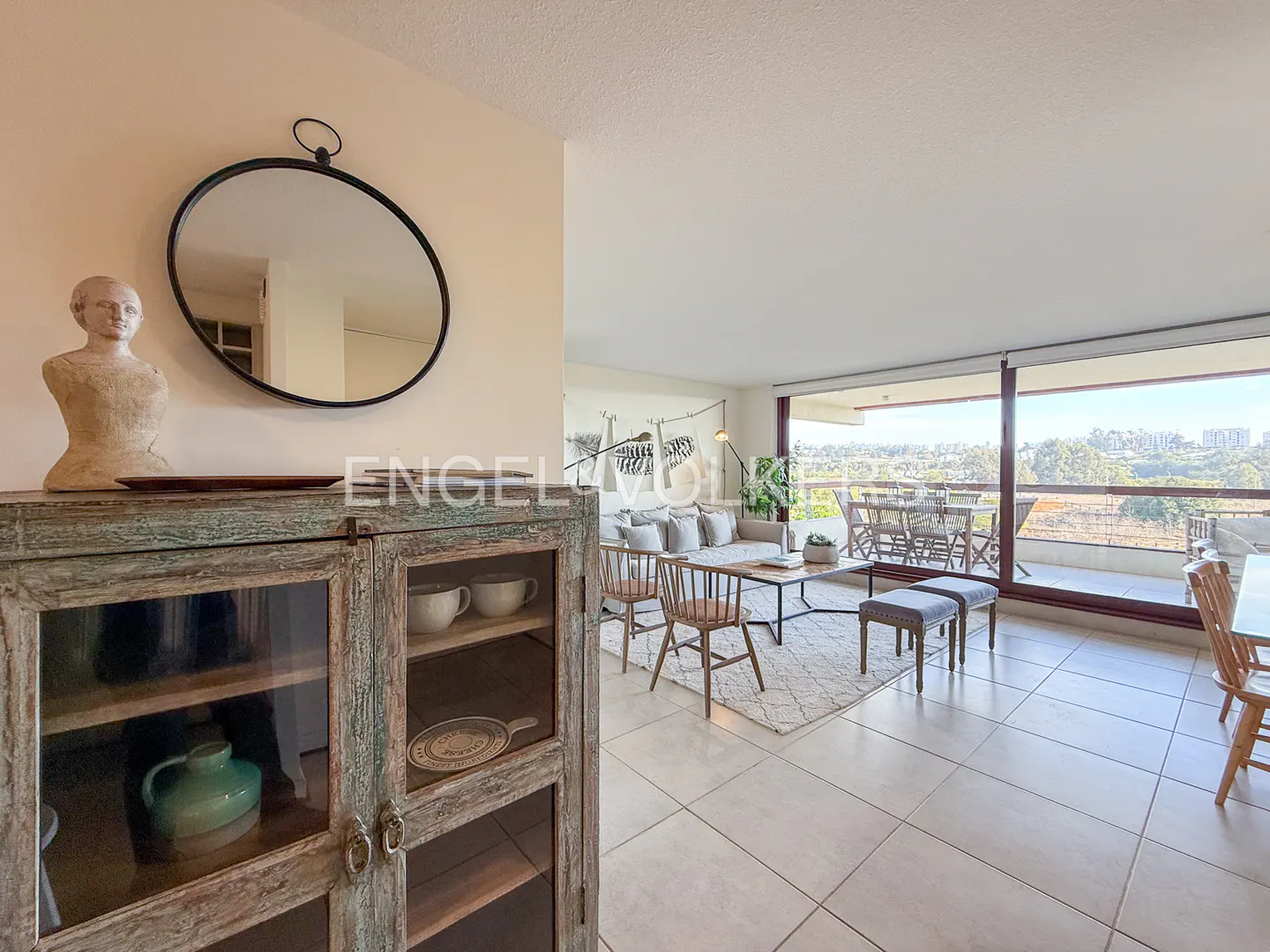 Living room with a rustic cabinet, bust, and round mirror. A sofa, chairs, and a balcony with a view are visible in the background.