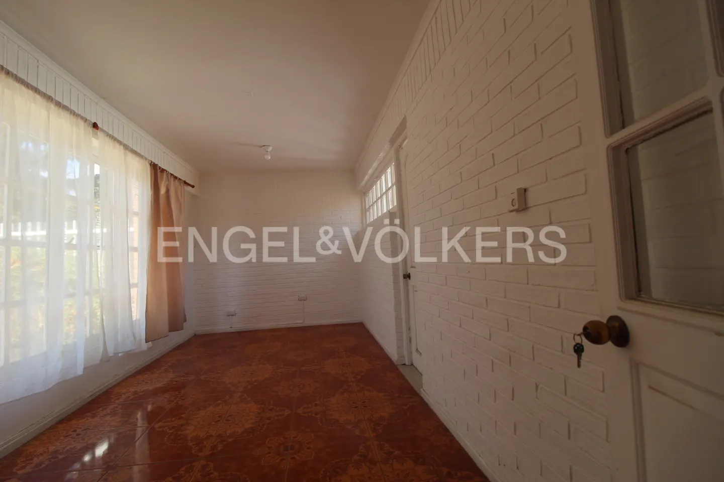 A bright room with white brick walls, a brown patterned floor, and sheer curtains on the window. A key hangs from the doorknob.