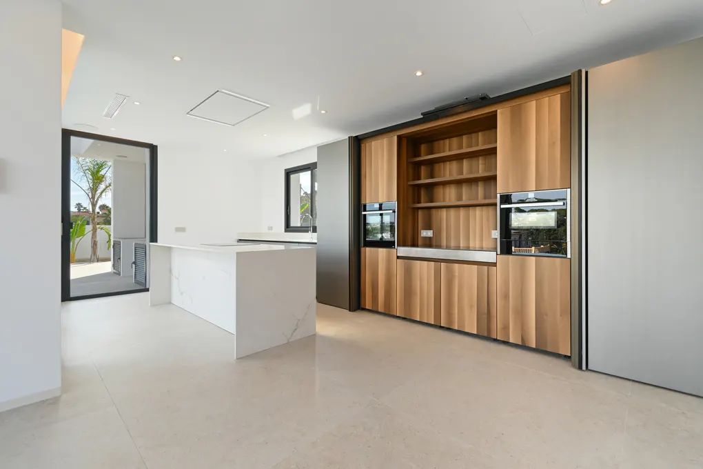 A modern, bright kitchen with white marble floors, a white island, and wood cabinets with built-in ovens. A black-framed glass door leads to an outdoor patio.