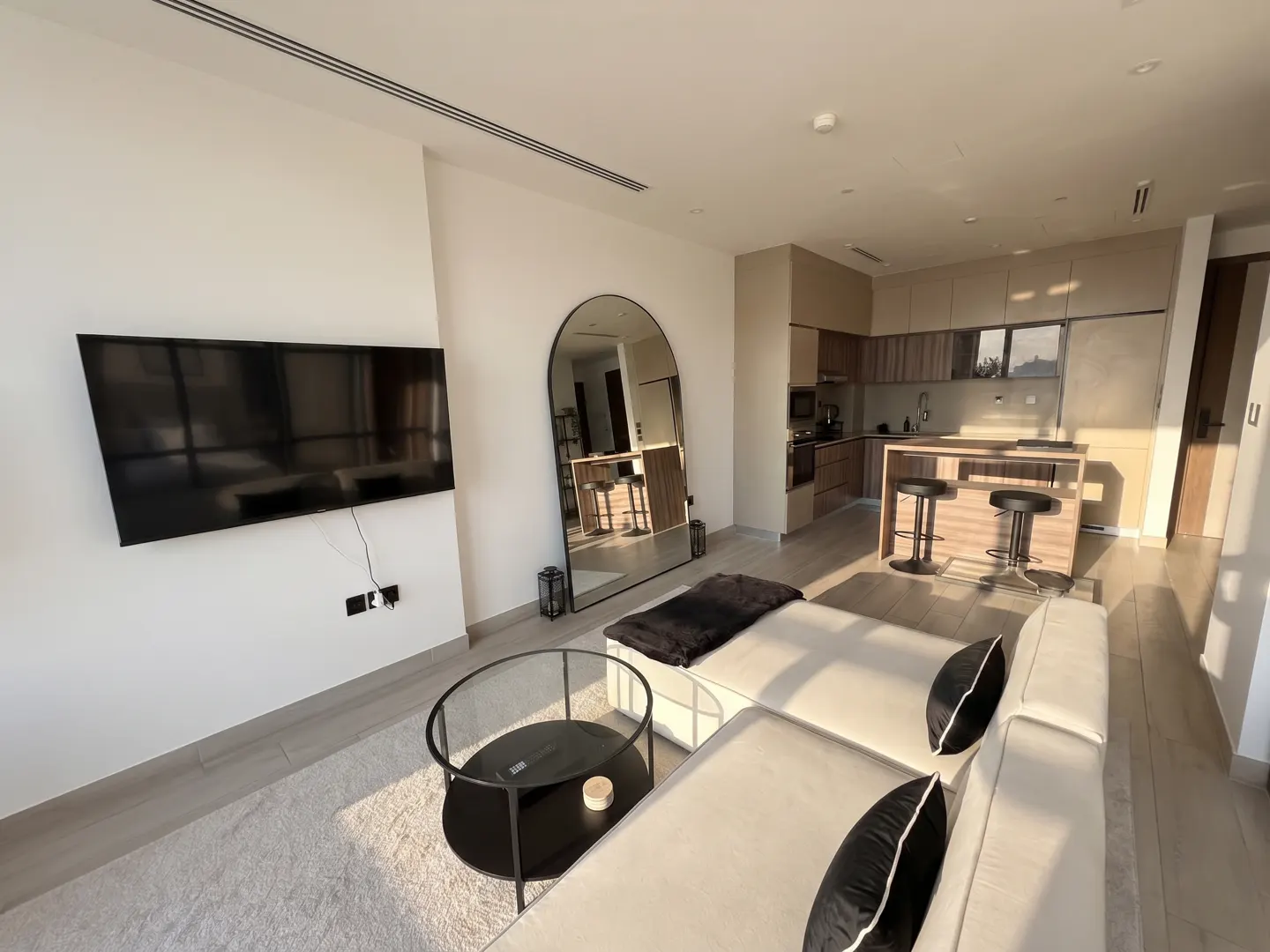 Bright, modern apartment interior with a white sofa, black pillows, and a glass coffee table. A large TV is mounted on the wall. The kitchen is visible in the background.