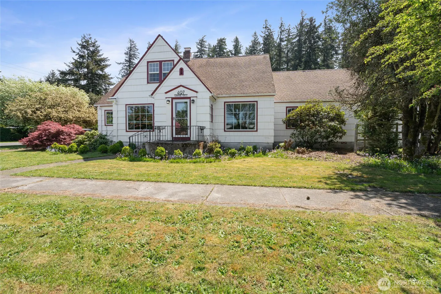 A white, one-story house with a brown roof and red trim, surrounded by a green lawn and trees.