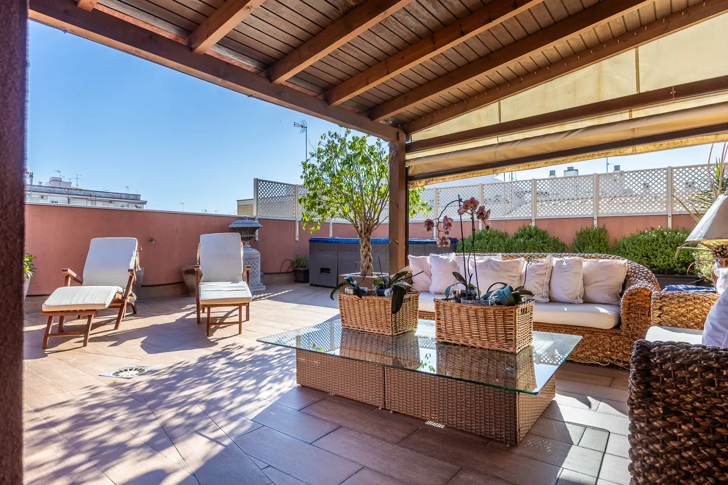 A sunny rooftop terrace with wicker furniture, lounge chairs, and a glass-topped coffee table. A wooden pergola provides shade.