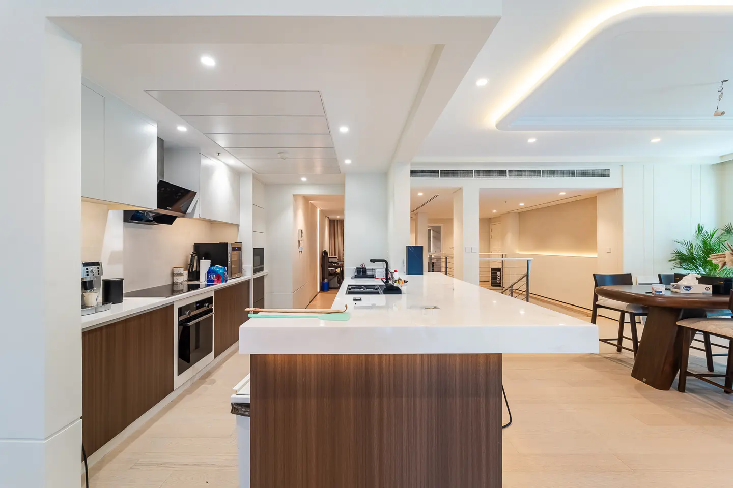 A modern kitchen with white cabinets, brown wood accents, and a white countertop island. A dining table and chairs are visible in the background.