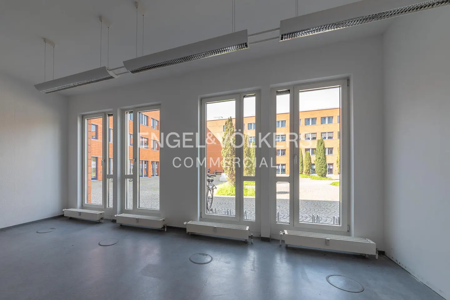 Bright, empty office space with white walls, grey floors, modern ceiling lights, and large windows overlooking a courtyard.