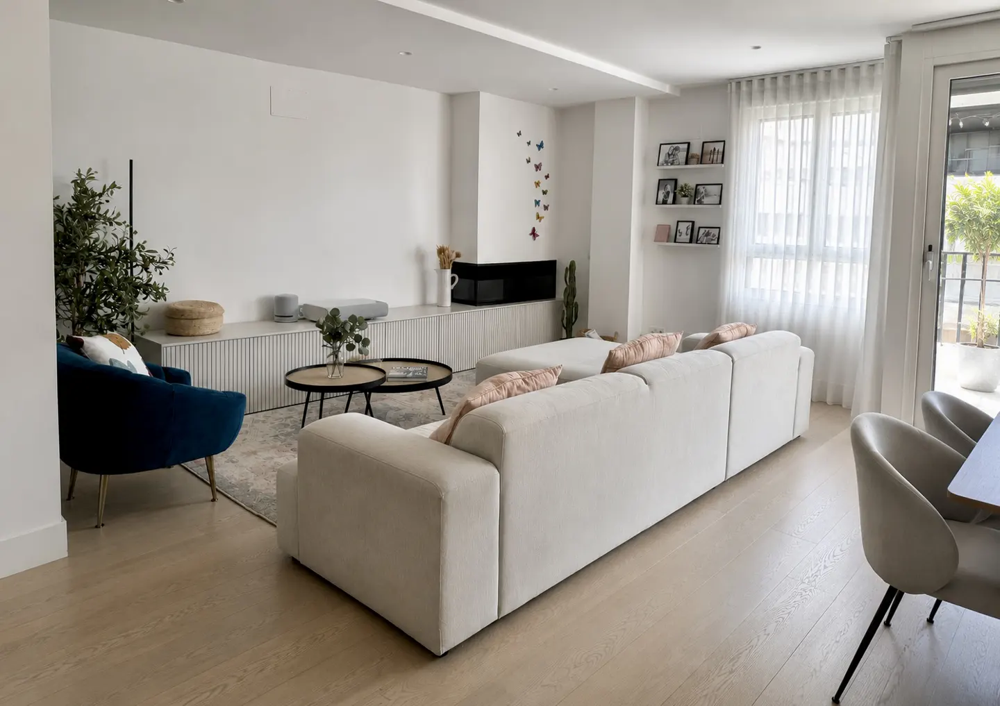 Bright living room with white walls, light wood floors, and a large white sofa with pink pillows. A blue chair and round tables sit near a built-in cabinet.