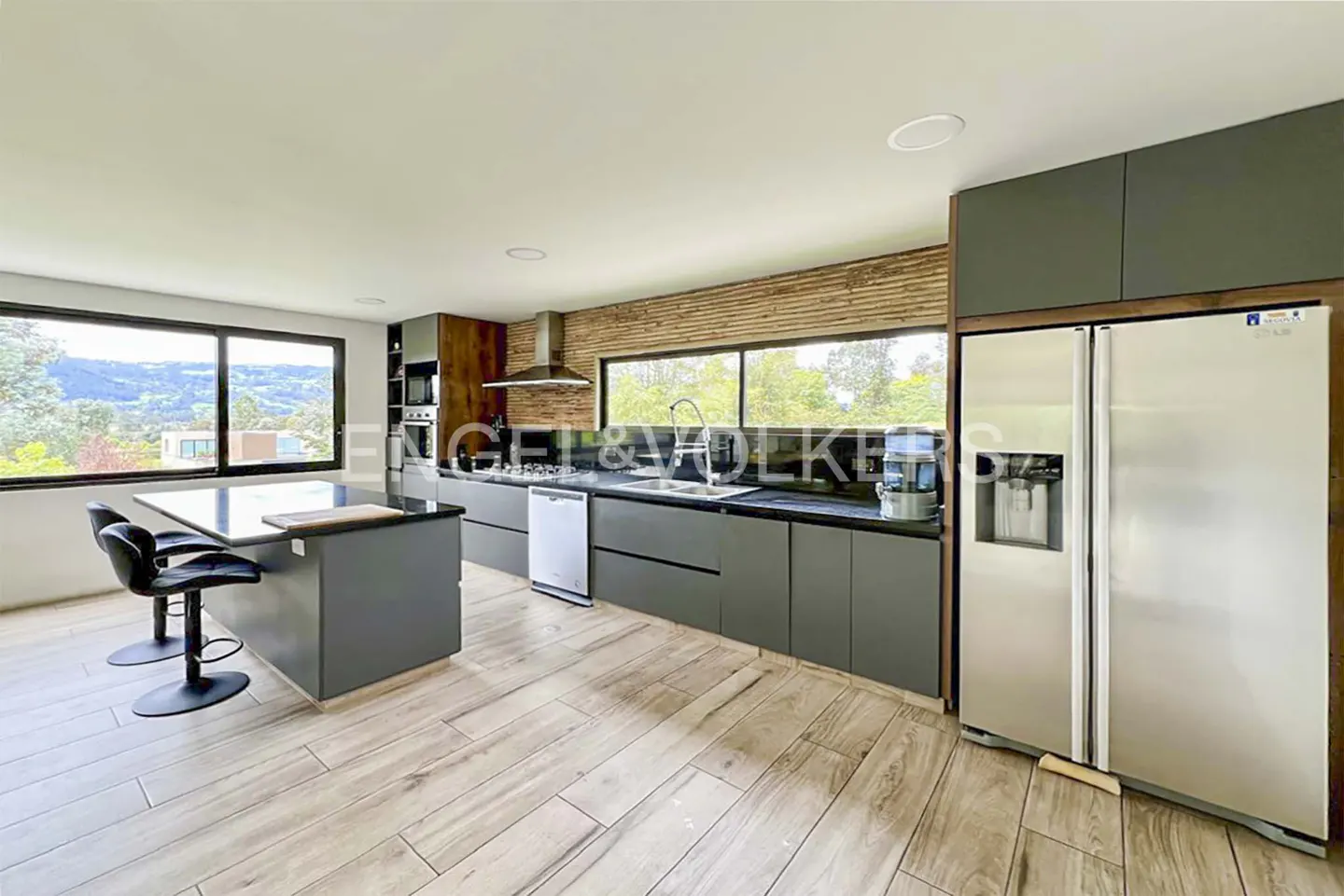 Modern kitchen with gray cabinets, stainless steel appliances, and wood-look tile flooring. Island with black countertop and two black stools. Large window with view.