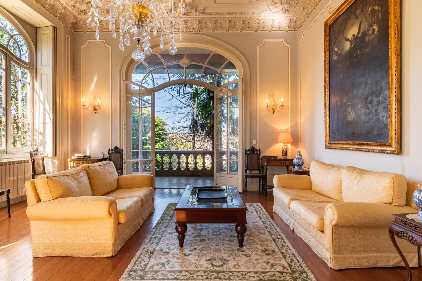 Elegant living room with yellow sofas, a chandelier, and an open door to a balcony with lush greenery. A large painting hangs on the wall.