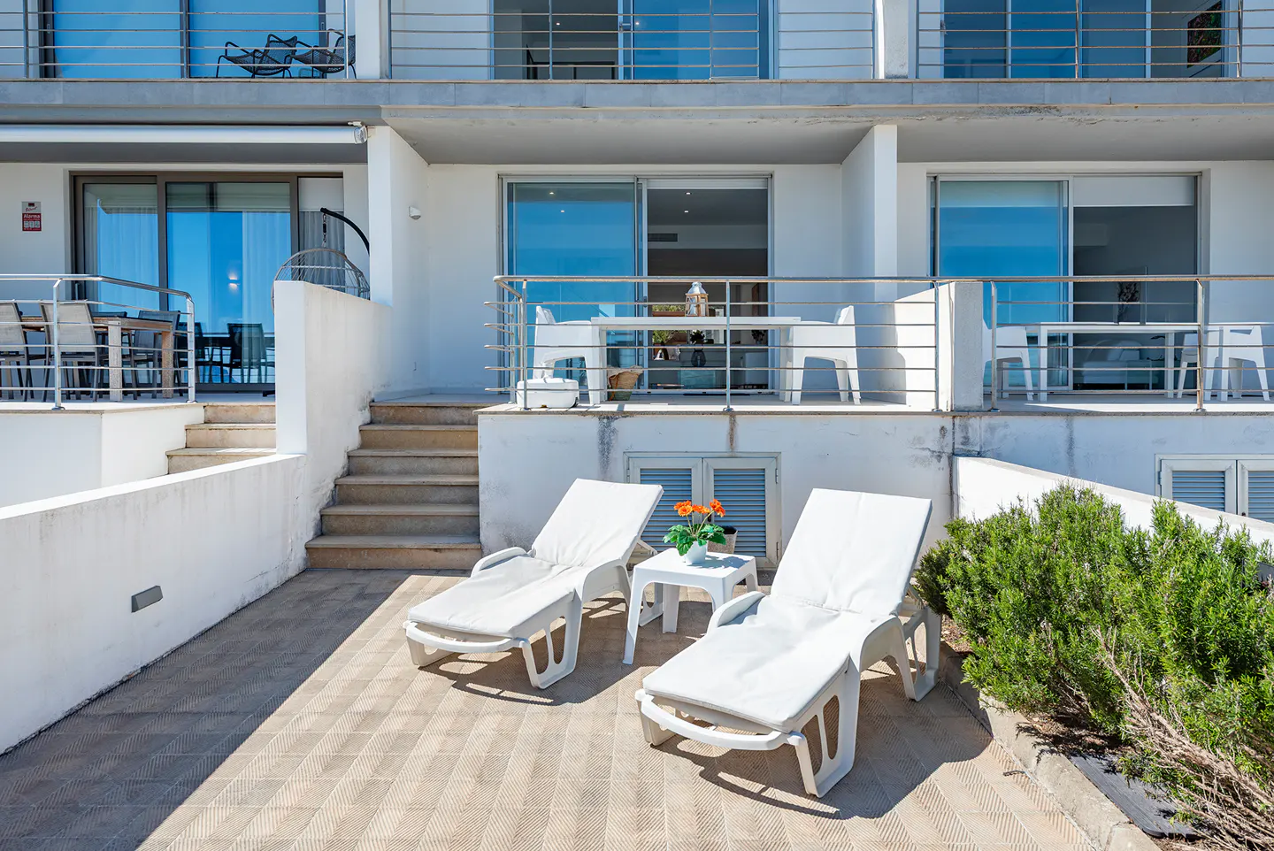 White building with balconies, stairs, and patio. Two white lounge chairs and a small table with flowers sit on the patio. Balconies have metal railings.