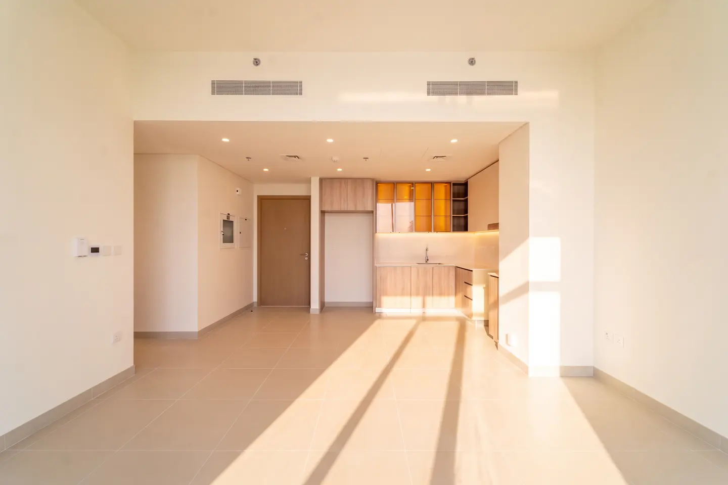 An empty apartment with tile floors, white walls, and a light wood kitchen. Sunlight streams in.
