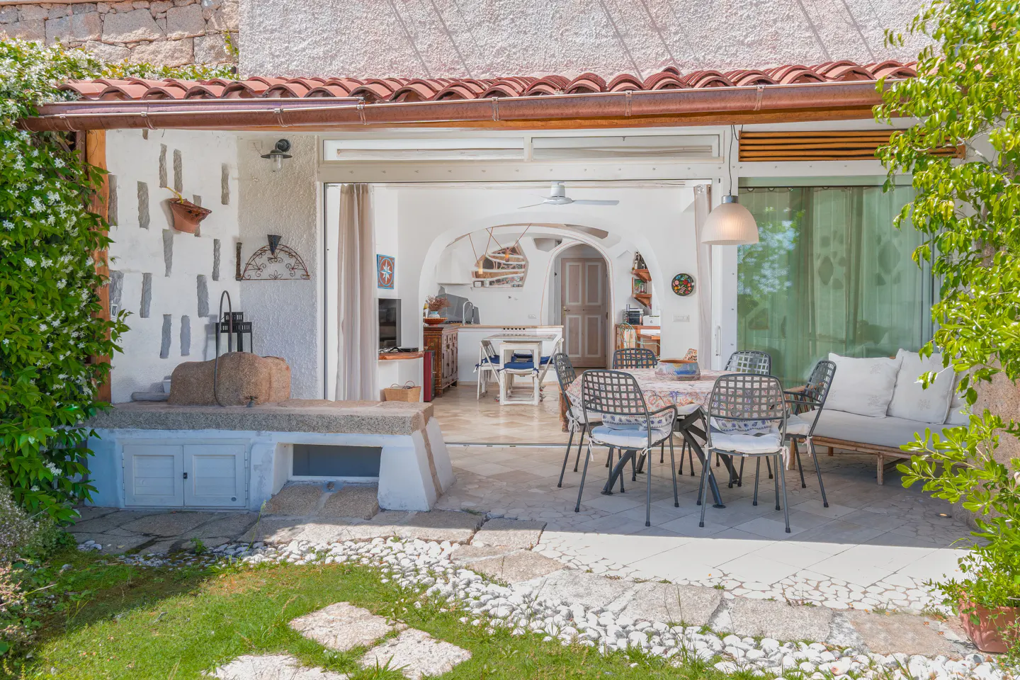 Covered patio with dining set, stone bench, and view into a bright, arched interior living space with white walls.