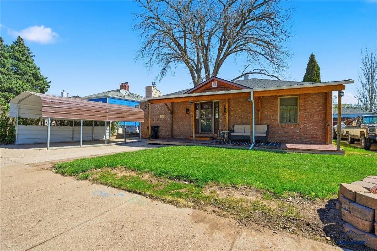 A single-story brick house with a covered porch, green lawn, and a metal carport on a sunny day.