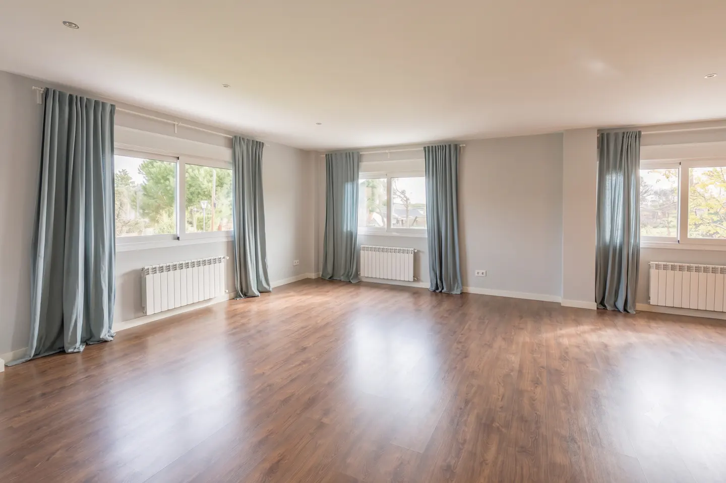 Spacious, empty room with light gray walls, dark wood floors, and large windows draped in soft blue curtains.