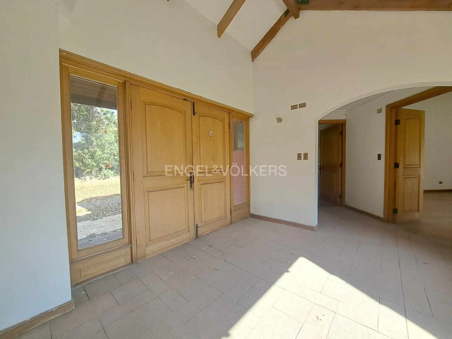 Interior view of a home's entrance with light wood double doors and tile flooring. Arched doorway leads to other rooms. Engel & Völkers logo on door.