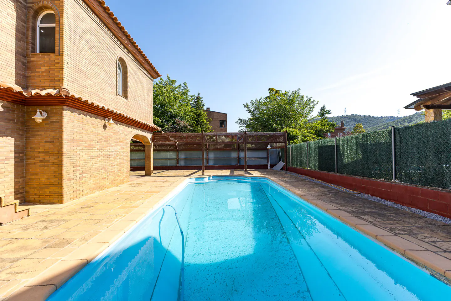A rectangular blue swimming pool is surrounded by a brick house and a green fence.