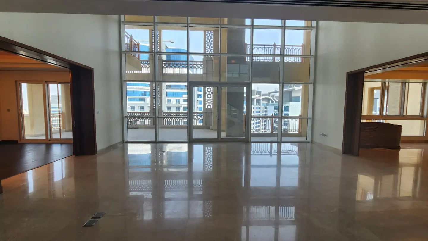 Empty living room with marble floors, large windows, and balcony access. Neutral walls and dark wood trim. Cityscape visible through the windows.