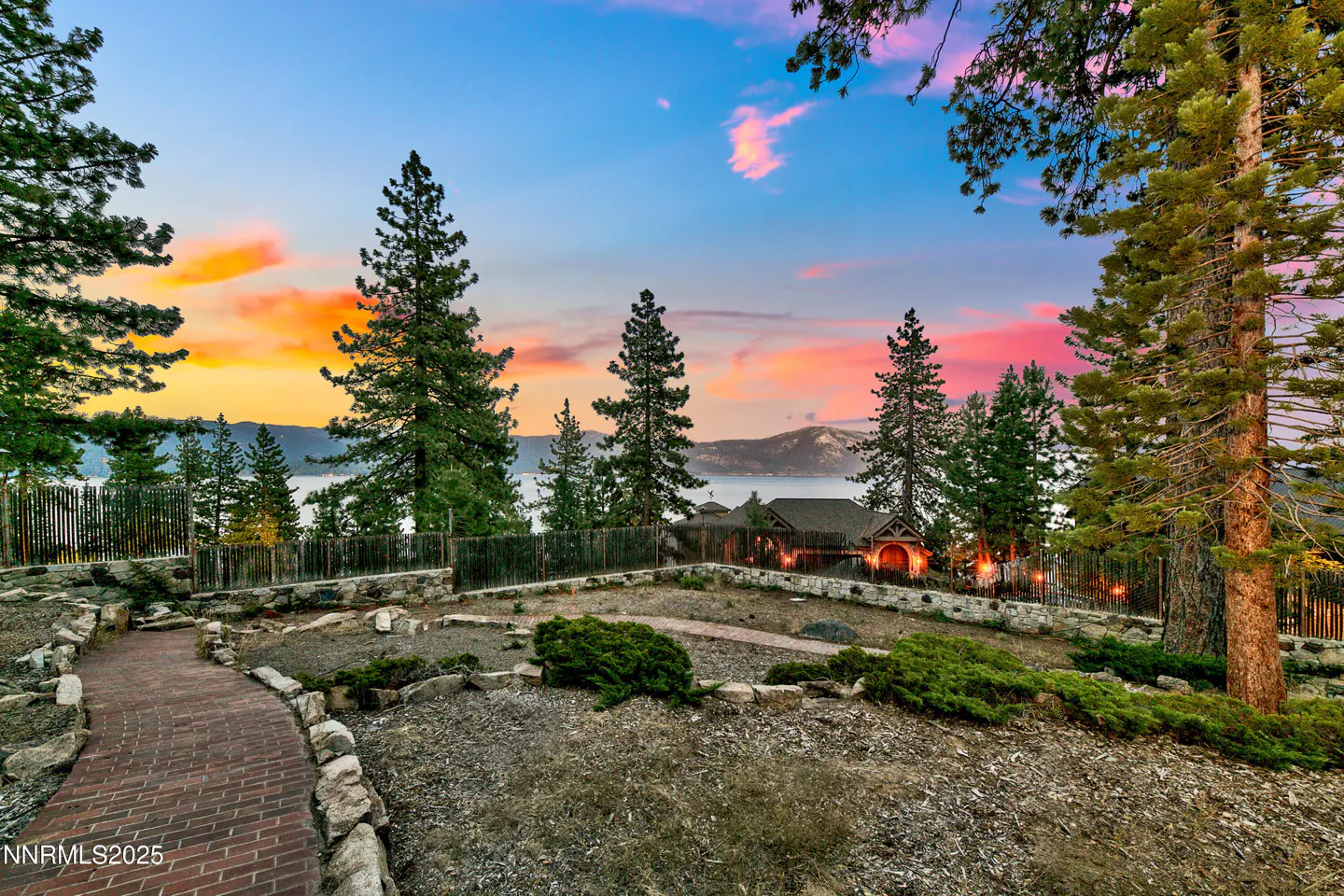 Scenic view of a property overlooking a lake at sunset. Tall trees frame a brick path leading to a house with a wooden fence.